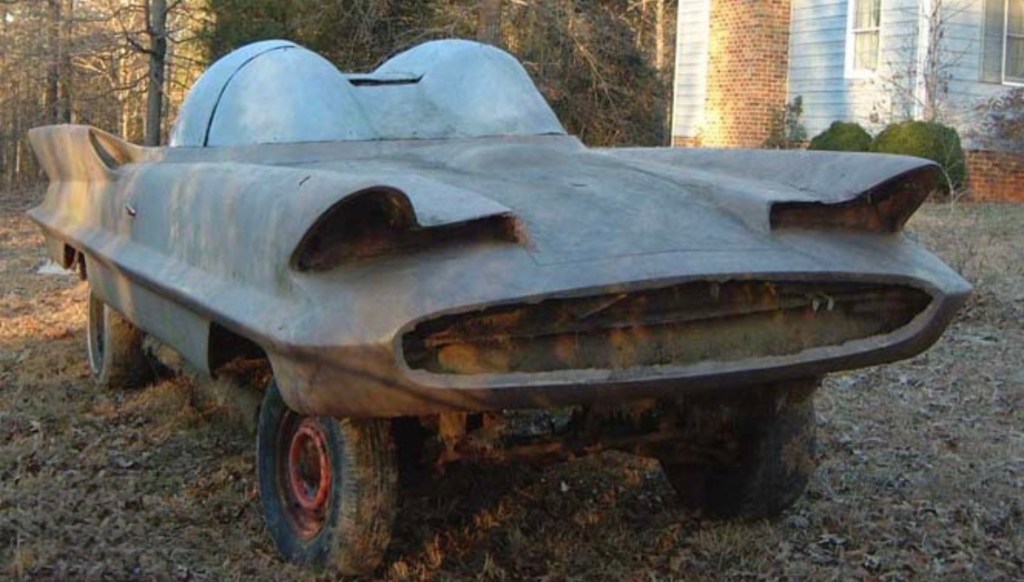 A vintage, rusty car with a unique, cartoonish design, featuring large headlights resembling eyes, parked in a rural area surrounded by trees.