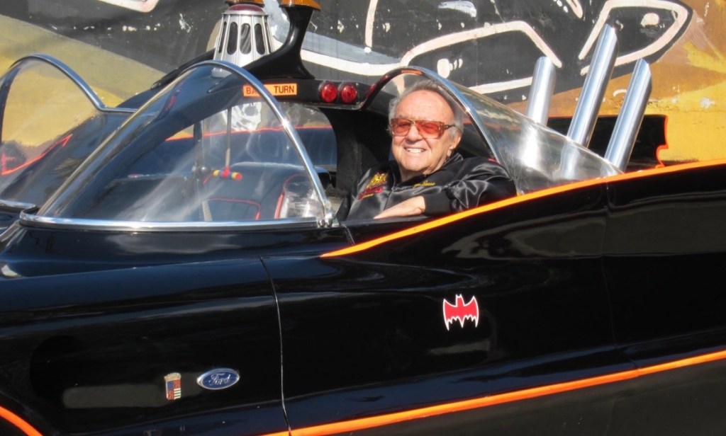 A smiling man wearing sunglasses sits inside a black car resembling the Batmobile, with a distinctive bat logo on the side.