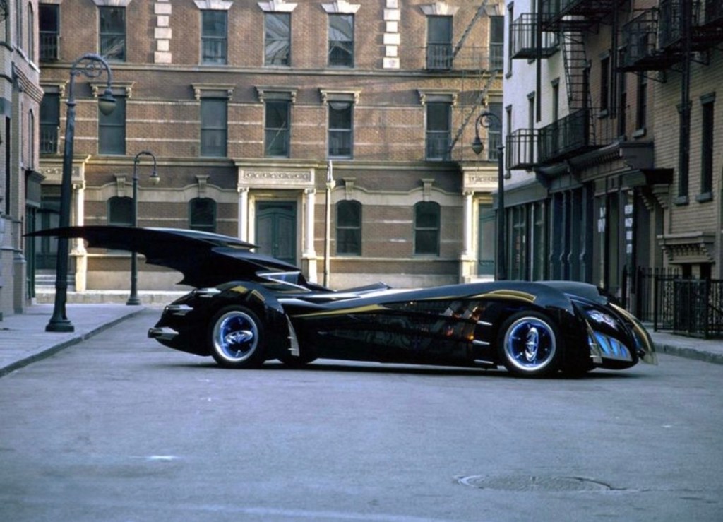 A sleek black car with a large wing parked on a deserted urban street, showcasing classic architecture in the background.