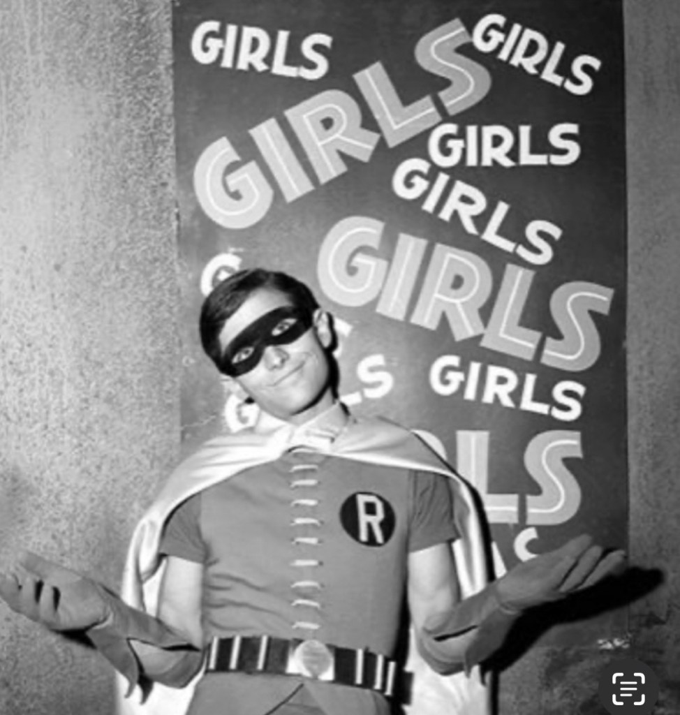 A young man dressed as a superhero in a costume with a cape and a mask, standing in front of a backdrop featuring repeated text that reads 'GIRLS'.