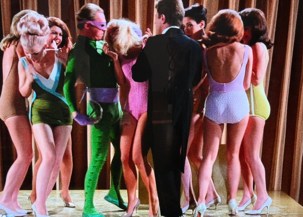 A group of women in colorful swimsuits surround a man in formal attire, creating a lively scene with a stage backdrop.