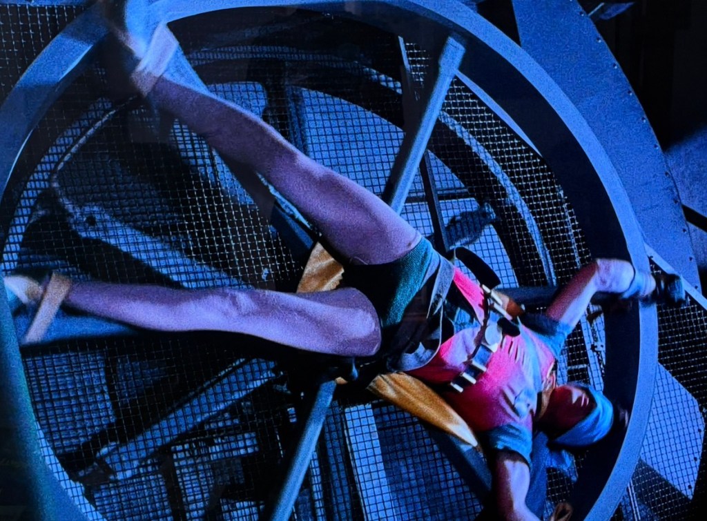 A person suspended in a harness, interacting with a large circular machinery component in a vivid blue setting.