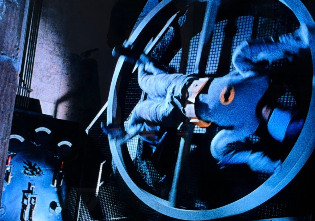 A person in a blue jumpsuit is training on a rotating apparatus, surrounded by control panels and machinery in a dimly lit environment.