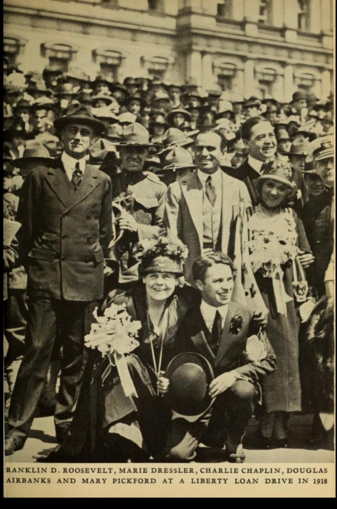 Historical gathering featuring Franklin D. Roosevelt, Marie Dressler, Charlie Chaplin, Douglas Fairbanks, and Mary Pickford at a Liberty Loan drive in 1918, surrounded by a crowd.