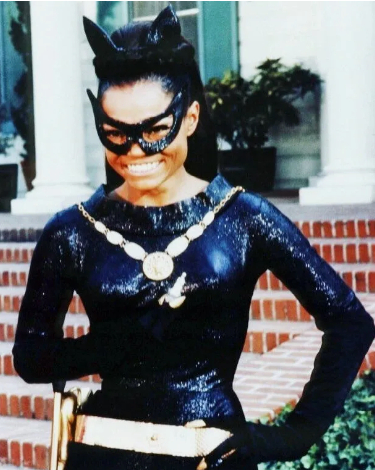 A woman dressed as a superheroine, wearing a black, shiny catsuit and a mask with cat ears, standing confidently with a smile. She has a gold belt and chain accessories, with a backdrop of stairs and greenery.