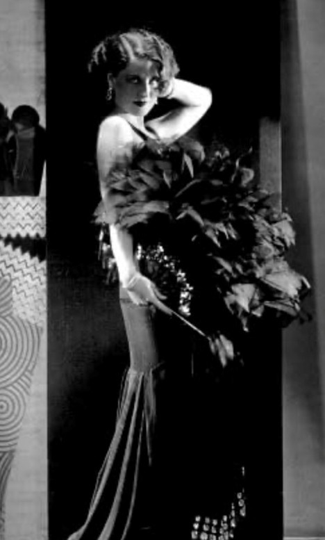 A glamorous woman in a vintage black dress poses elegantly with a large feather boa and a fan, showcasing a classic Hollywood style.