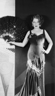 A glamorous woman in a stylish, form-fitting evening gown poses elegantly with a fan, set against a backdrop featuring artistic patterns.