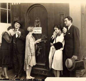 A group of five people in early 20th-century attire, standing in front of a closed movie theater door with a 'CLOSED' sign. They appear to be upset or disappointed, holding ice cream cones.