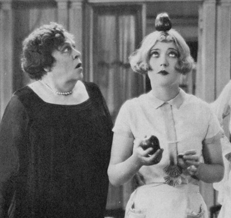 A woman in a black dress looks surprised while another woman in a short white dress balances an apple on her head and holds another apple in her hand.