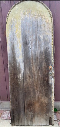 Weathered wooden door with an arched top, featuring a rusty latch and intricate metal detailing.