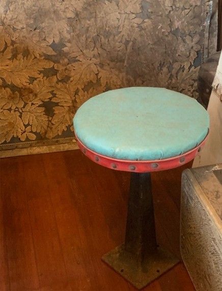 A vintage-style stool with a turquoise padded seat and a red rim, supported by a black metal base, placed against a decorative wallpaper with leaf patterns.