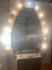 A large, illuminated mirror surrounded by lights, with a vintage suitcase placed in front.
