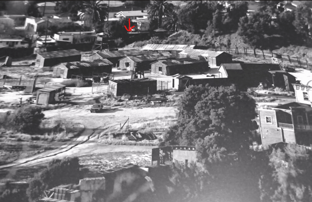 Aerial view of a small, rustic settlement featuring wooden shacks, dirt roads, and sparse vegetation, with a few palm trees in the background and a prominently marked building indicated by a red arrow.