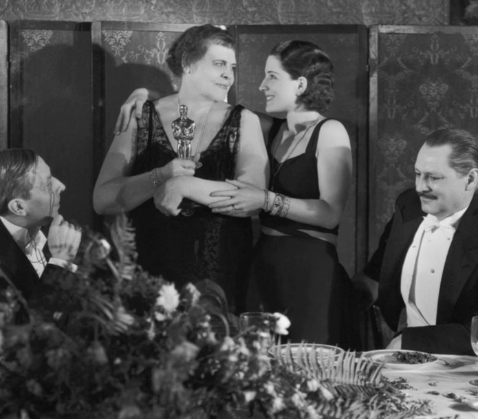 A historic black and white photo of four individuals at a banquet, two women embracing with an Oscar trophy, while two men watch, one dressed in a tuxedo.