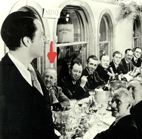 A black and white photo of a formal dinner setting with several men seated at a long table, focusing on a well-dressed speaker standing at the front, addressing the group.
