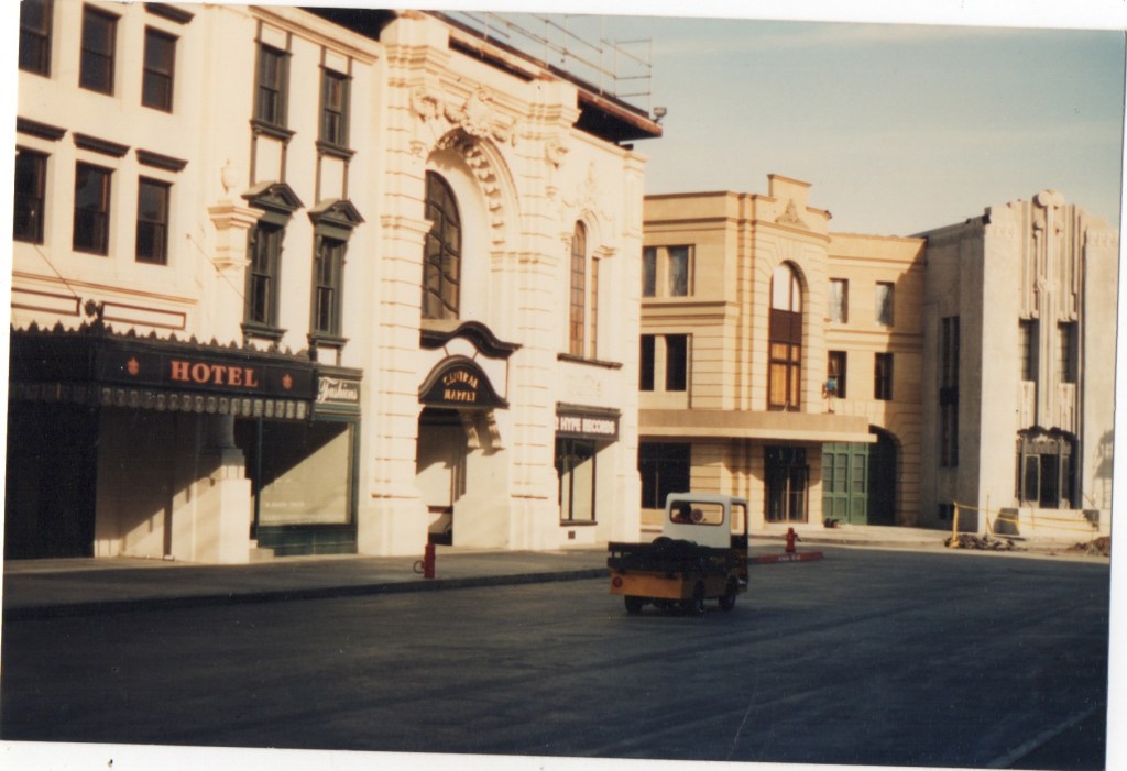 A view of a film studio street scene featuring buildings with various architectural styles, including a hotel and a theater, with a small vehicle driving down the empty street.