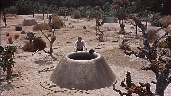 A man in a white shirt examines a large, dome-shaped structure in a barren landscape, surrounded by unusual vegetation and other similar structures.