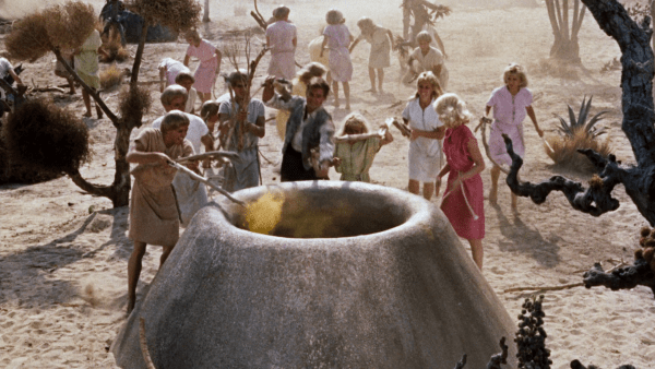 A group of people gathering around a large, round cauldron in a desert setting, stirring a mixture while others watch attentively.