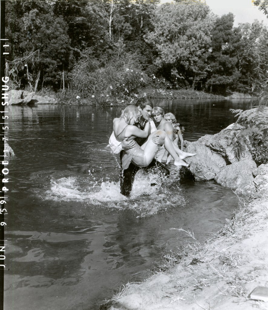 A black and white photo of four people playing by a river. A man is lifting two women while another woman stands nearby, all enjoying a sunny day outdoors.
