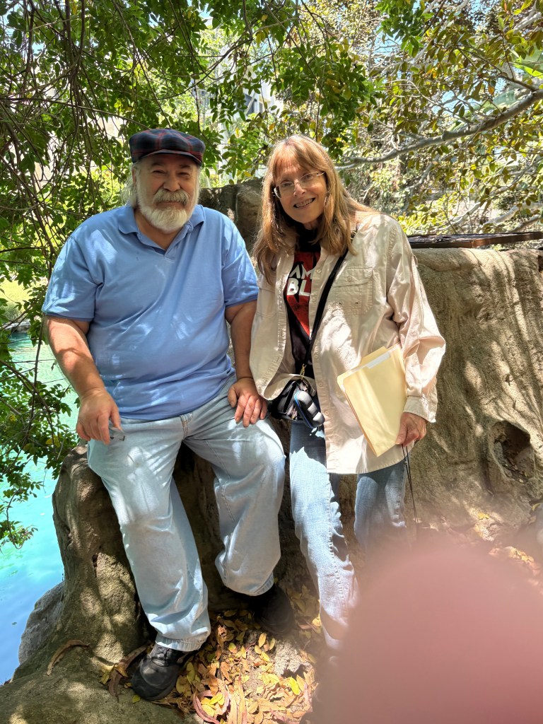 A man with a beard wearing a blue shirt and hat sits beside a woman in a light jacket, both smiling while sitting on a large rock surrounded by greenery.