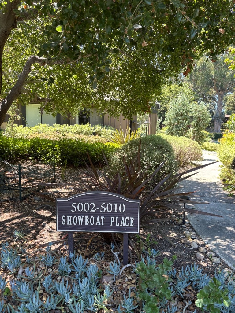 A wooden sign displaying '5002-5010 SHOWBOAT PLACE' surrounded by lush greenery and garden plants.