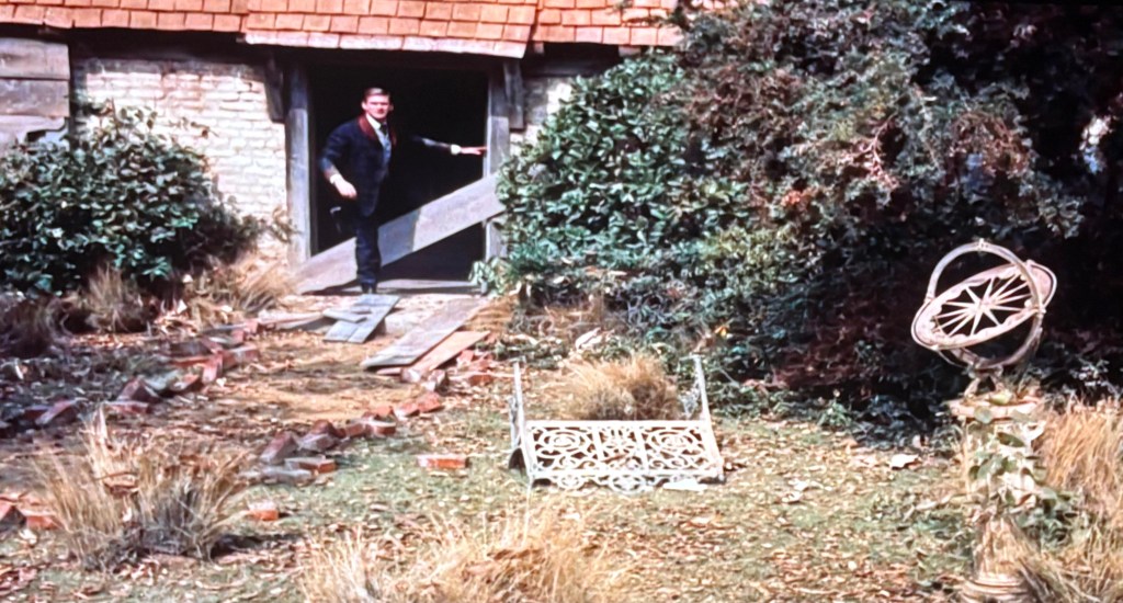 A man standing at the entrance of a barn, gesturing with his hand. The area features overgrown grass and bushes, with a decorative garden feature visible in the foreground.