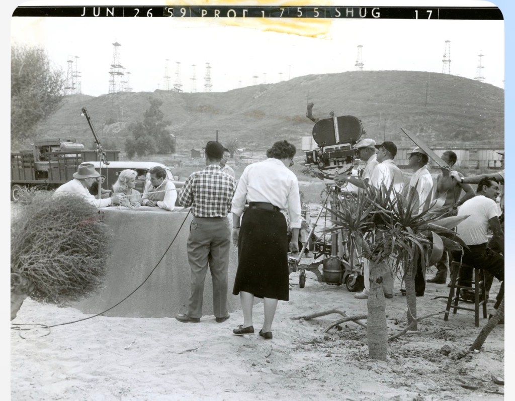 Black and white photograph of a film crew on location, featuring a table discussion among actors, a large camera setup, and various crew members in a sandy landscape with oil rigs in the background.