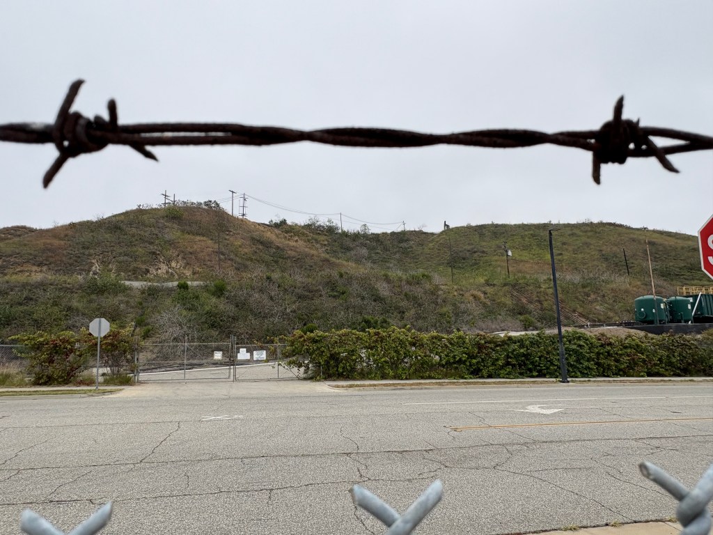 A barbed wire fence in the foreground with a hilly landscape and power lines in the background, under a cloudy sky.