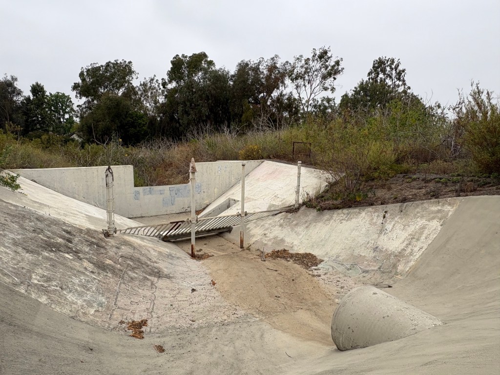 An abandoned concrete drainage area surrounded by overgrown vegetation, featuring a dilapidated wooden bridge and a large cylindrical concrete structure.
