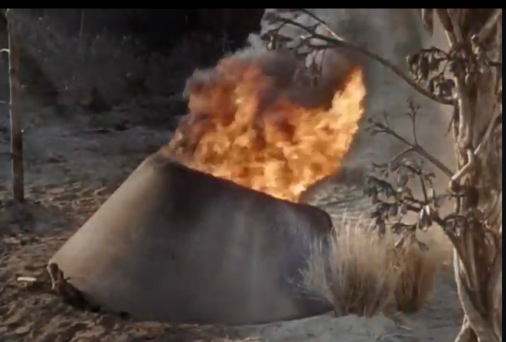 A large metal object emitting flames in a barren landscape, with sparse vegetation around.