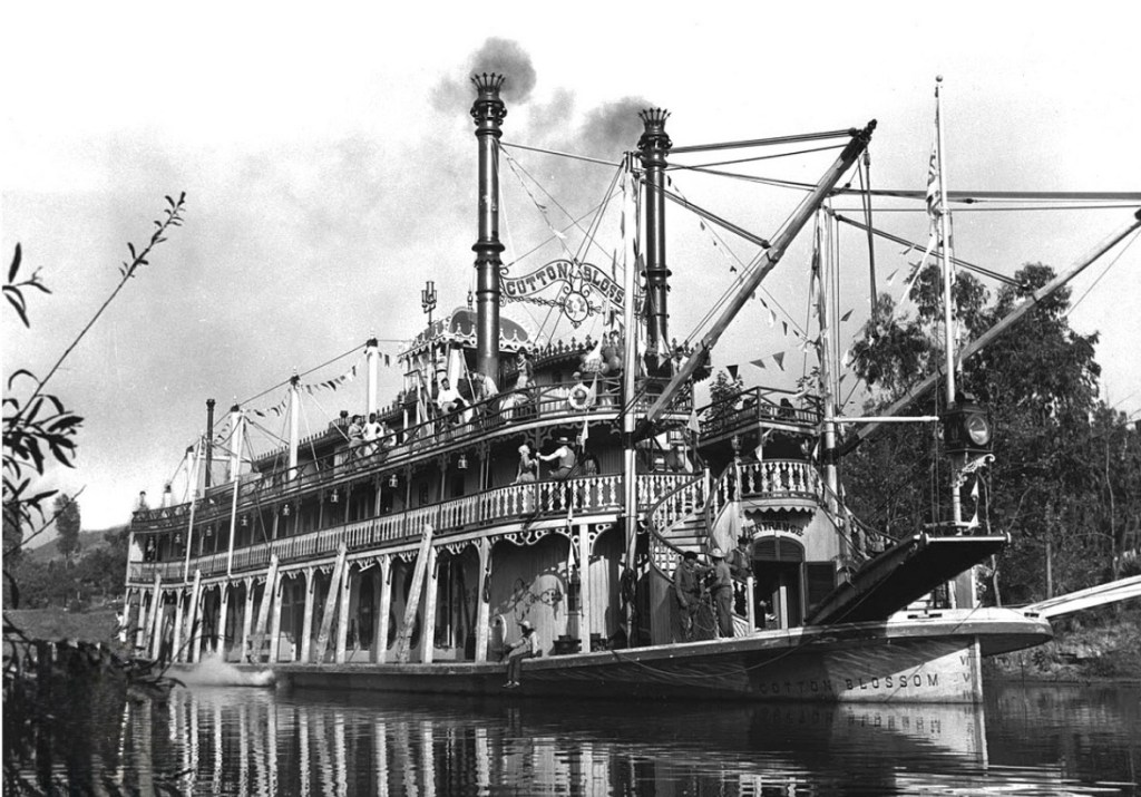 A historic steamboat named 'Cotton Blossom' with multiple decks and smokestacks navigating a river, decorated with flags and a few people aboard.