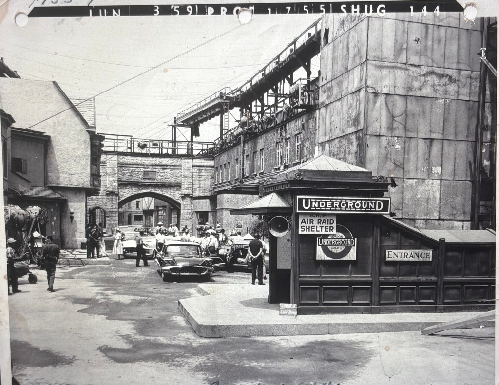 A vintage black-and-white photograph of a street scene, featuring an underground entrance marked as 'UNDERGROUND' and 'AIR RAID SHELTER.' Several classic cars are parked nearby, with people walking and standing around, set against a backdrop of buildings and structures.