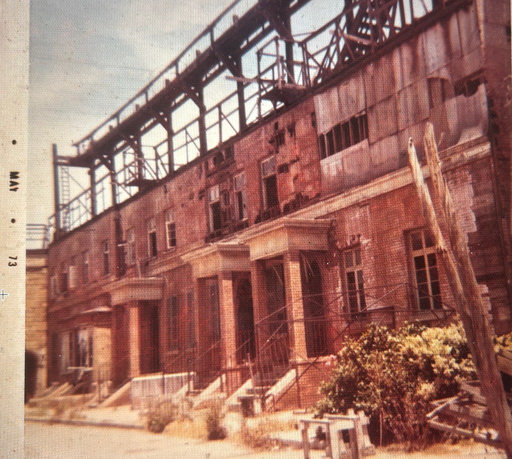 Abandoned building with peeling paint and broken windows, featuring a decaying facade and visible structural damage, surrounded by overgrown vegetation.