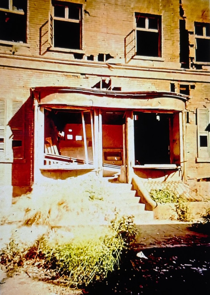 A close-up view of an abandoned building's entrance, featuring broken windows and dilapidated steps surrounded by overgrown vegetation.