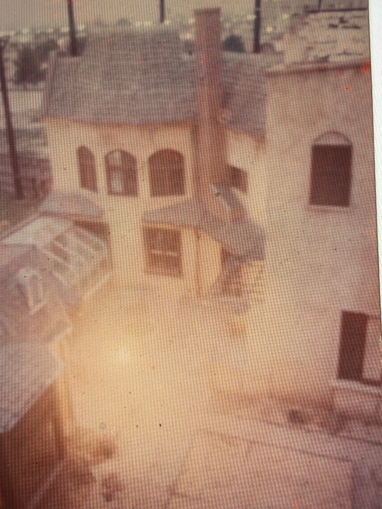An aerial view of a vintage house with a sloped roof, featuring multiple windows and a central chimney, surrounded by empty outdoor space.