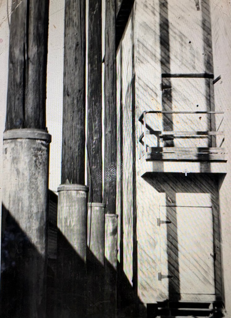 Black and white photograph featuring several vertical wooden posts and a structure with a door and railing, casting shadows on the nearby surface.