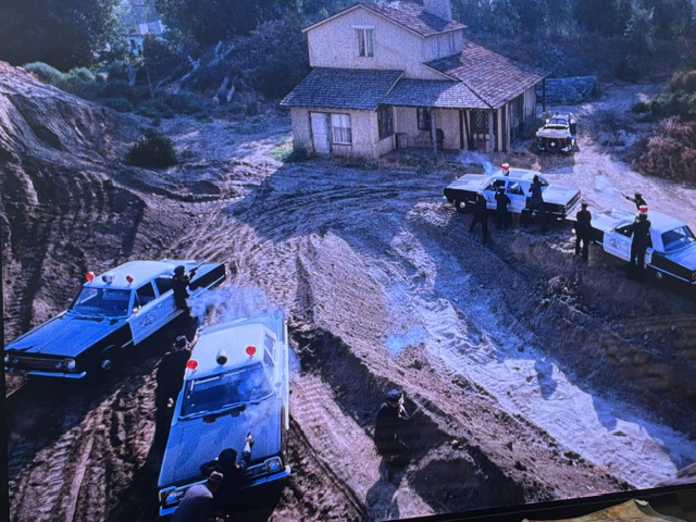 Aerial view of several police cars with lights flashing near a house on a dirt road, with officers on the ground in a tense situation.