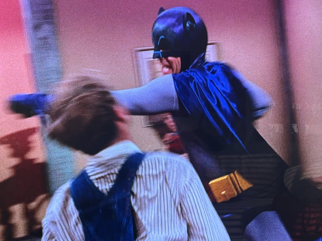 A classic scene depicting a superhero in a bat costume throwing a punch at a character wearing overalls in a colorful room.
