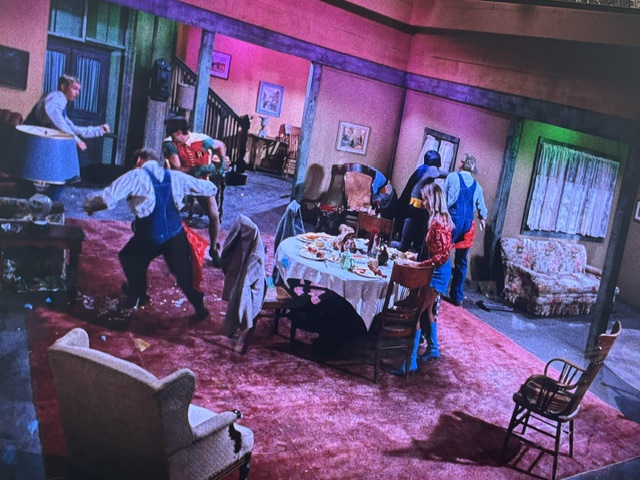 A chaotic room with several people involved in a lively activity, featuring a dining table with leftover food and drinks, a couch, and colorful lighting.