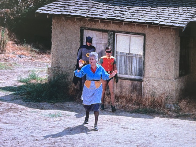 A scene featuring a woman in a blue dress with a yellow apron, standing in front of a small house, while two men in superhero costumes, one in gray and the other in red and green, are positioned behind her.
