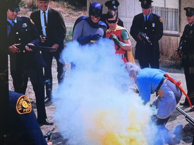 Batman and Robin in costumes, surrounded by police officers, as colorful smoke billows from an object being handled by a person on the ground.
