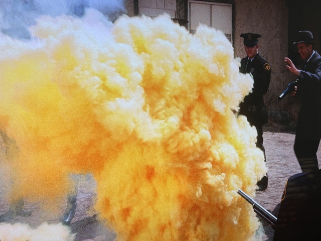 A scene depicting a large cloud of yellow smoke, with two police officers in uniform standing nearby, one holding a firearm.