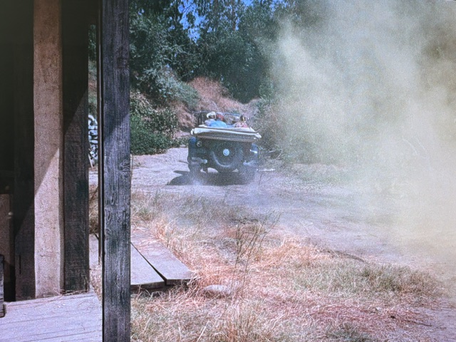 A vintage car driving on a dusty road surrounded by dry grass and vegetation, partially obscured by dust.