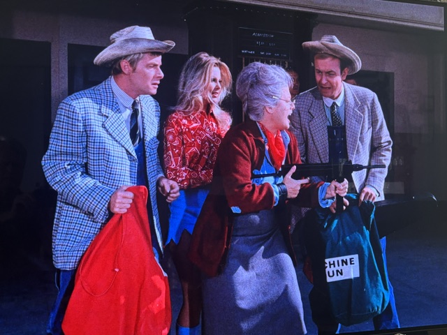 A group of four characters, two men and two women, dressed in colorful outfits and hats, interact humorously. One of the women holds a toy gun, while they carry bags with various colors.