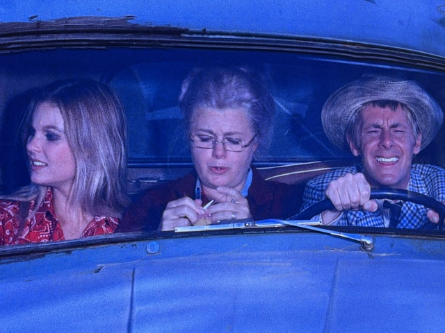 A comedic scene featuring three people in a car, with a woman in a red patterned shirt on the left, an older woman with glasses in the middle, and a man driving, looking stressed.