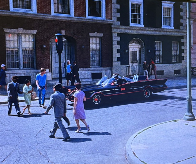 A classic black car reminiscent of the Batmobile is stopped at a traffic light on a busy street, while pedestrians cross and walk along the sidewalk in a lively urban setting.