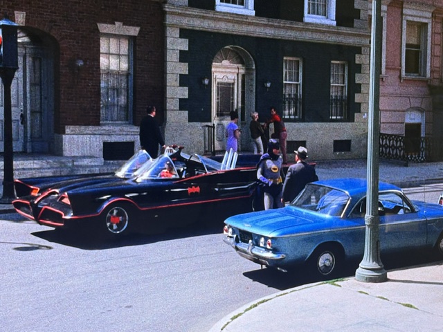 A vintage scene featuring the iconic Batmobile and a blue car on a city street. Several individuals, including a man in a costume, are gathered near a building as they interact. The streets are lively with pedestrians and a traffic light visible.