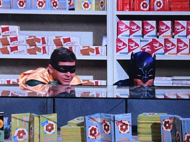 Two masked superheroes, Robin and Batman, peering over a display case filled with colorful snack boxes in a store.