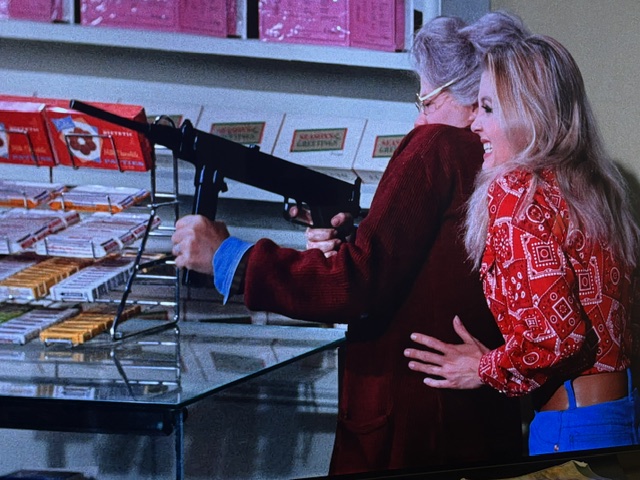 A scene featuring a person holding a firearm while another person stands beside them, both in a retail setting with display racks of products in the background.