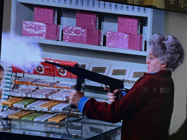 A woman in a red sweater is aiming an assault rifle in a store with cigarette displays, smoke visible from the gun.